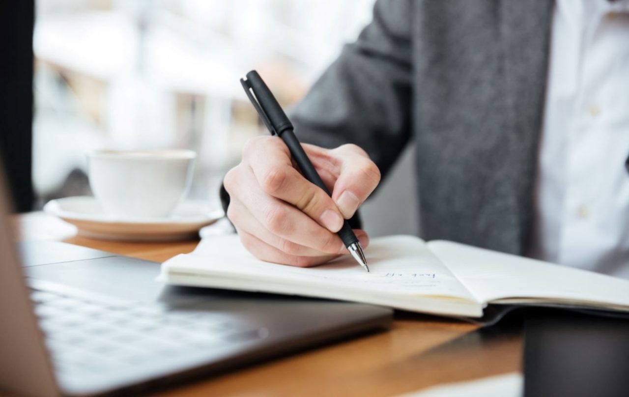 imagem-recortada-de-empresario-sentado-junto-a-mesa-de-cafe-com-computador-portatil-e-escrever-algo imagem-recortada-de-empresario-sentado-junto-a-mesa-de-cafe-com-computador-portatil-e-escrever-algo