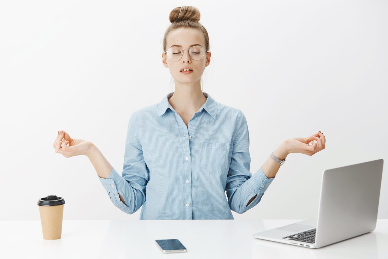 Girl meditating at work sitting in lotus pose with nirvana orbs near laptop and smartphone trying relax and get rid of pressure and stress posing calm and peaceful against gray background in glasses